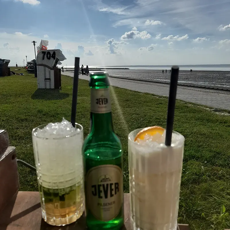 Eine Jever Flasche steht zwischen zwei Cocktails auf einem Tisch. Dahinter sind Strand, Meer und Strandkörbe.