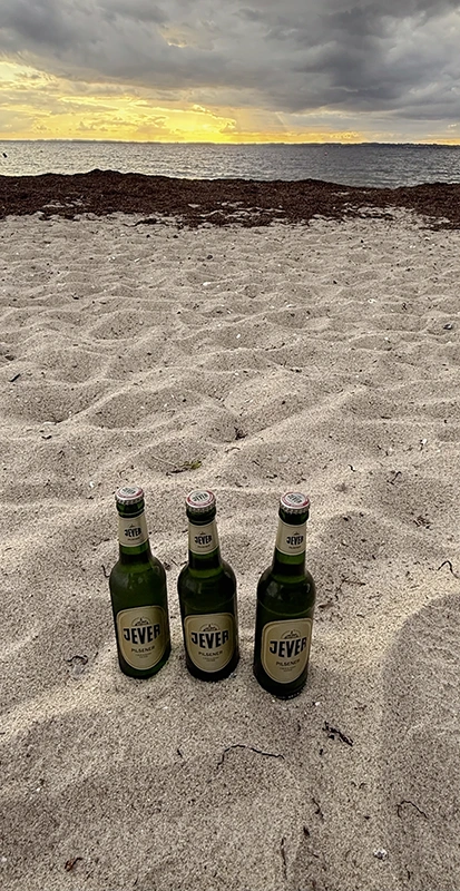 Drei Jever Bierflaschen stehen auf sandigem Strand. Dahinter dunkle Wolken und ein Sonnenuntergang über dem Meer.