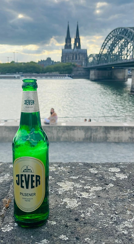 Grüne Jever-Flasche auf grauem Steg vor Kölner Dom und Hohenzollernbrücke bei bewölktem Himmel.