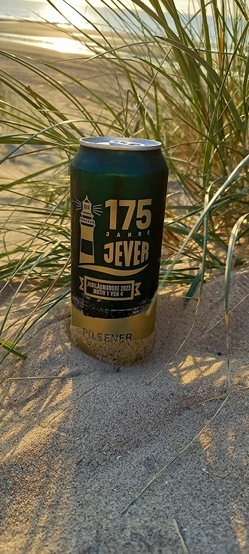 Grüne Jever-Dose mit 175-Jahre-Logo im Sand vor Dünengras, verschwommener Strand und Meer im Hintergrund
