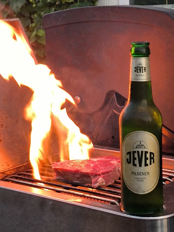 Jever-Flasche steht neben einem Stück Fleisch auf einem heißen Grill mit lodernden Flammen im Hintergrund.