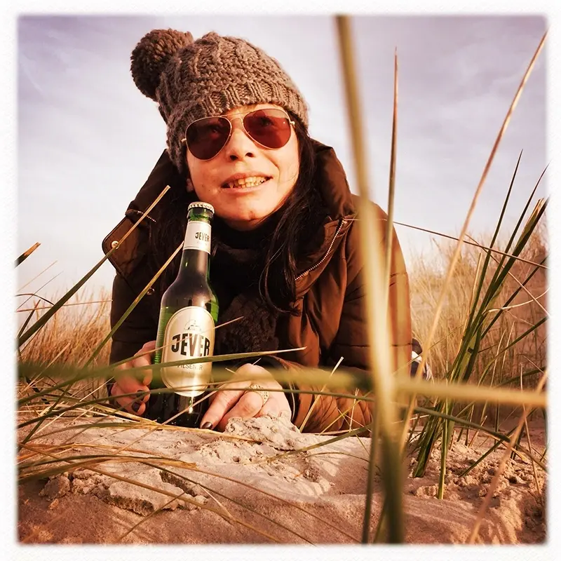 Frau mit Jever Bierflasche im Strandgras. Blick in die Kamera.