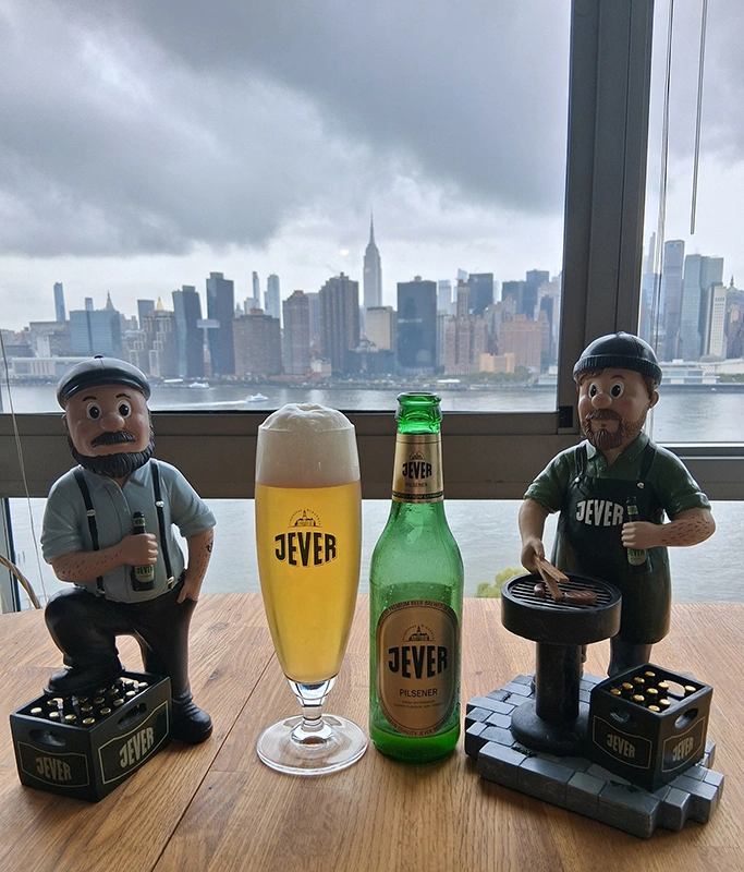 Zwei Figuren stehen auf einem Tisch, dazwischen eine Flasche Jever und ein gefülltes Glas, vor der Skyline von New York. 