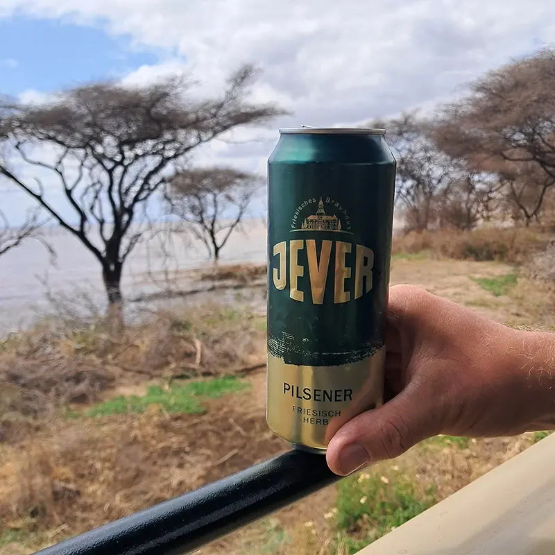 Eine Jever Pilsener Dose wird von einer Han auf einem Geländer gehalten. Im Hintergrund sind ein See und Bäume.