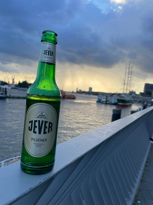 Eine Jever Flasche steht auf einem grauen Metallgeländer am Hafen. Im Hintergrund bewölkter Himmel und Boote im Wasser.