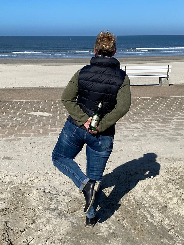 Person steht am Strand auf einem Fuß und hält eine Jever Bierflasche hinter dem Rücken. Ansicht von hinten.