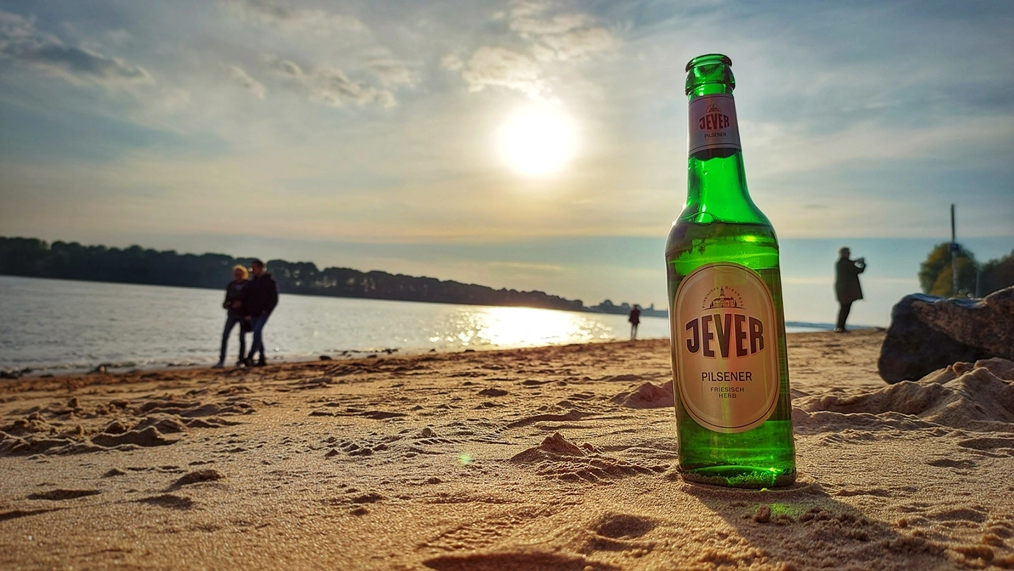 Eine Jever-Bierflasche steht bei einem Strand im Sand. Im Hintergrund befinden sich mehrere Personen.