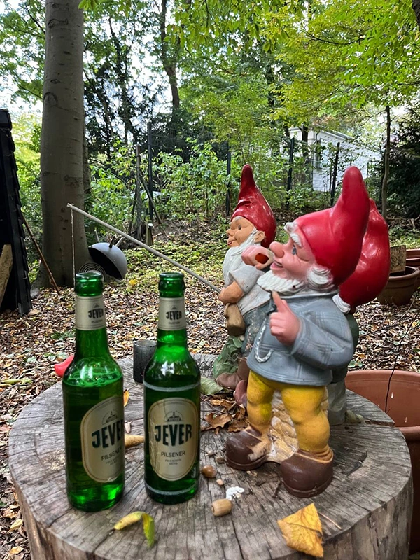 Auf einem Baumstumpf stehen zwei Jever-Bierflaschen. Rechts neben ihnen stehen drei Gartenzwerge. Einer davon angelt in Richtung der Flaschen.