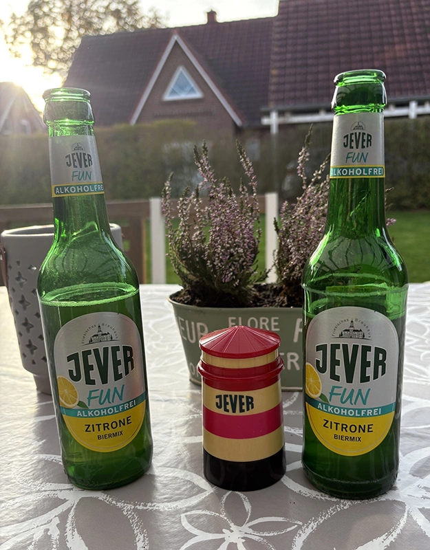 Auf einem Tisch in einem Garten stehen zwei Jever-Bierflaschen. Zwischen den beiden Flaschen steht ein kleiner Leuchtturm mit dem Schriftzug Jever.