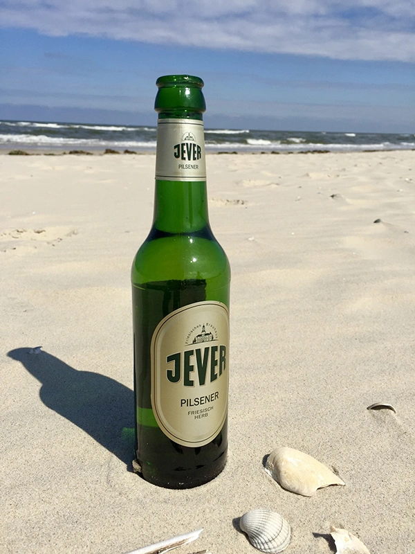 Eine Jever-Bierflasche steht an einem Strand im Sand. Neben ihr liegen Muscheln und im Hintergrund brechen Wellen.