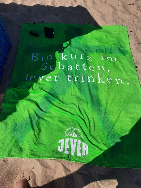 Ein grünes Handtuch mit dem Aufdruck Bin kurz im Schatten Jever trinken und dem Jever-Logo darunter liegt an einem Sandstrand.