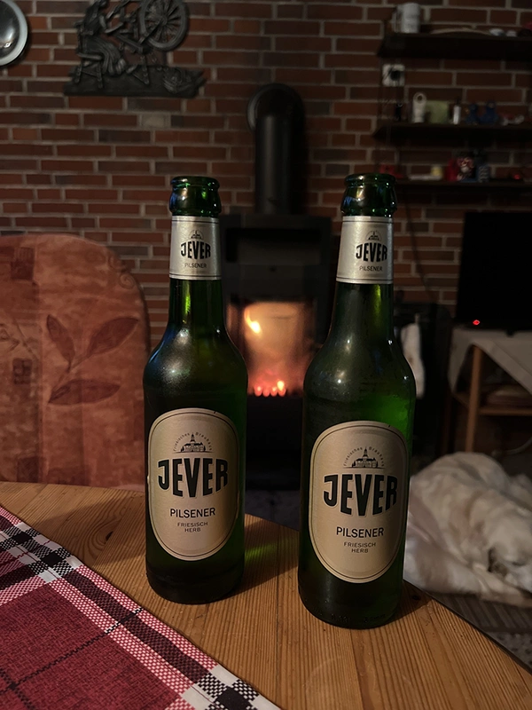 Zwei Jever-Bierflaschen stehen auf einem Holztisch nebeneinander. Im Hintergrund liegt ein Hund schlafend vor einem Kaminofen, in dem ein Feuer brennt.