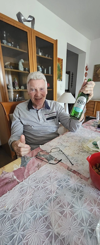 Ein Mann sitzt in einem Wohnzimmer an einem Tisch und hält eine Bierflasche der Marke Jever in der Hand.
