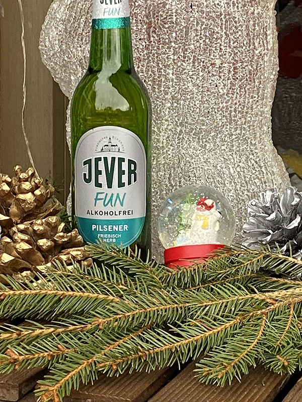 Eine Jever-Bierflasche steht links neben einer Weihnachtsschneekugel. Links und rechts von ihr liegt jeweils ein Tannenzapfen. Vor der Flasche liegt ein Tannenzweig.