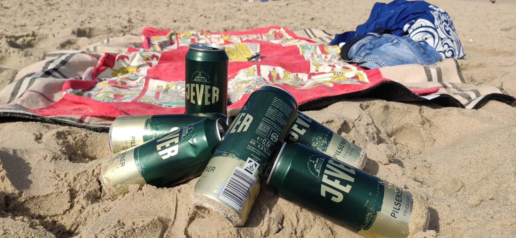 Mehrere Jever-Bierflaschen liegen aneinander gelehnt im Sand. Im Hintergrund liegt ein Handtuch mit Kleidung.