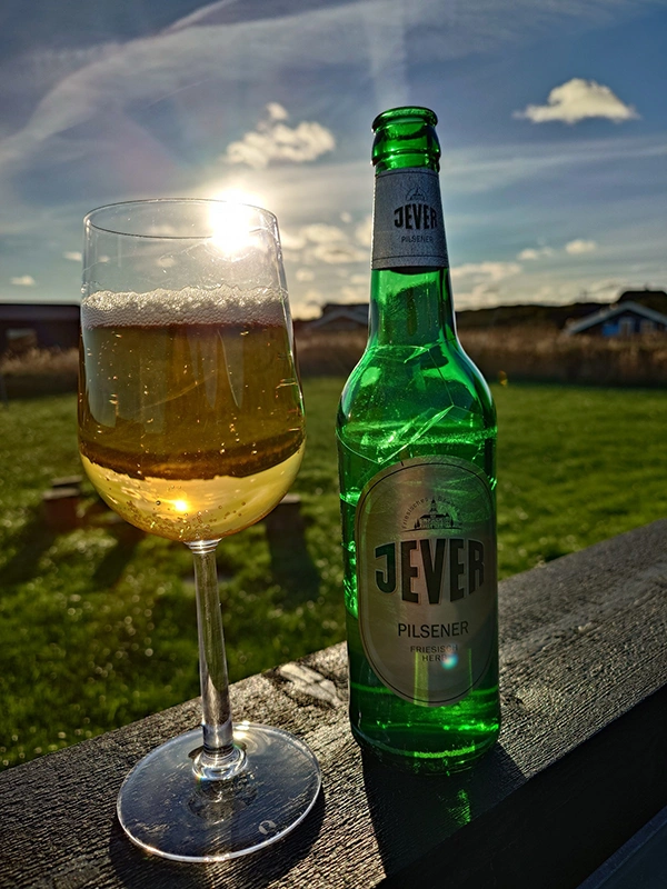 Eine Jever-Bierflasche steht auf einem Balken. Links neben ihr steht ein Glas mit Bier darin. Im Hintergrund ist eine Wiese mit einem Haus.