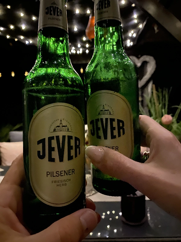 Zwei Hände halten Jever-Bierflaschen nebeneinander. Im Hintergund sind viele Lichter.