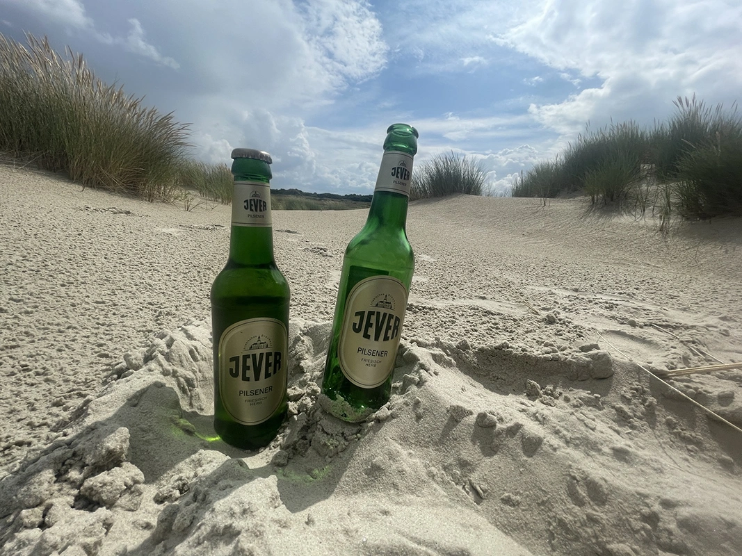 Zwei Jever-Bierflaschen stehen im Sand. Im Hintergrund sind Dünen mit Gestrüpp.