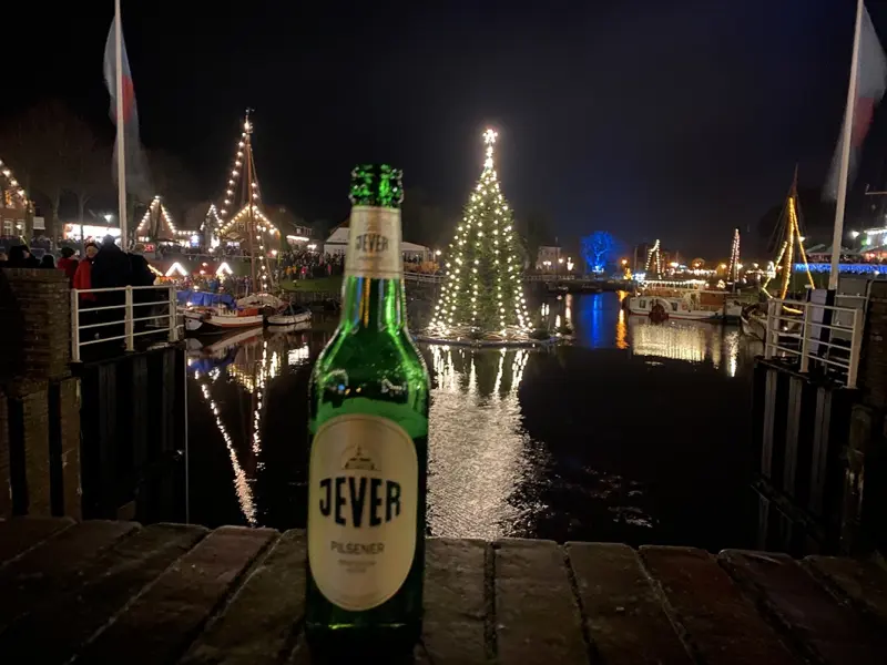 Eine Jever-Bierflasche steht auf einer Mauer. Im Hintergrund steht ein beleuchteter Tannenbaum in einem Gewässer. Um das Wasser herum stehen mehrere beleuchtete Hütten.