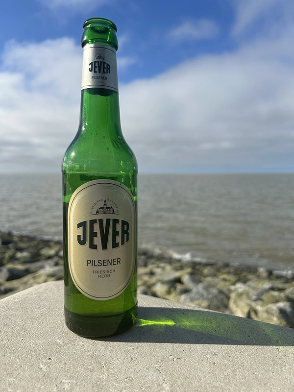 Eine Jever-Bierflasche steht am Rand einer Steinmauer. Im Hintergrund schwappt ein Gewässer gegen Steine.