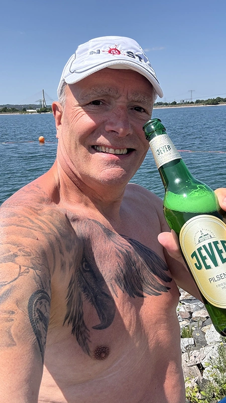 Ein Mann, der vor einem Gewässer steht, hält eine Jever-Bierflasche neben sein Gesicht.