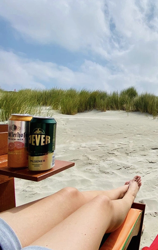 Eine Person liegt in Blickrichtung zu Dünen auf einer Liege im Sand. Links neben ihr steht eine Jever- und eine Schöfferhofer-Bierflasche.