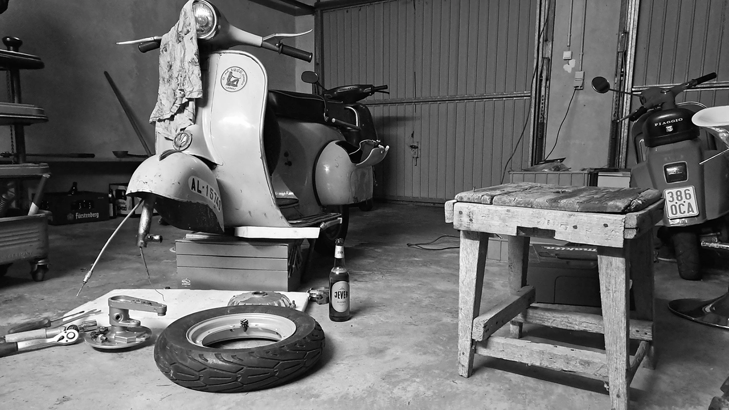 Eine Jever-Bierflasche steht auf dem Boden einer Garage. Um sie herum sind Fahrzeuge, Fahrzeugteile und Werkzeug.