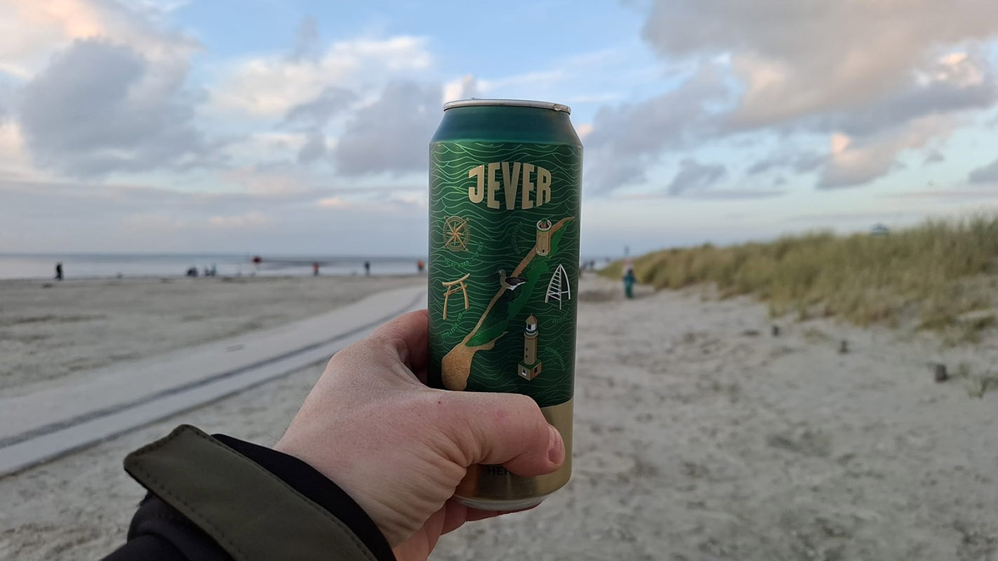 Eine Person hält eine Jever-Bierdose hoch. Im Hintergrund ist ein Strand. Rechts sind Stranddünen.