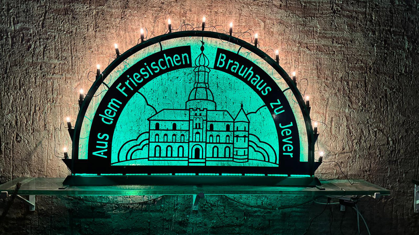 Ein Schwibbogen mit der Aufschrift Aus dem Friesischen Brauhaus zu Jever steht auf einem Wandregal. Er wird grün beleuchtet.