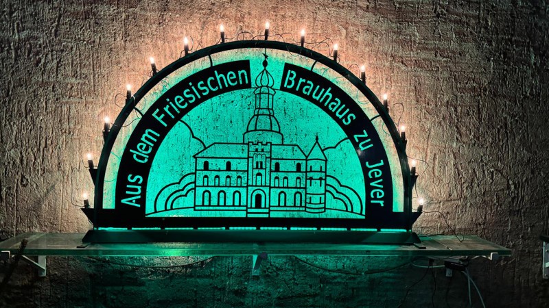 Ein Schwibbogen mit der Aufschrift Aus dem Friesischen Brauhaus zu Jever steht auf einem Wandregal. Er wird grün beleuchtet.