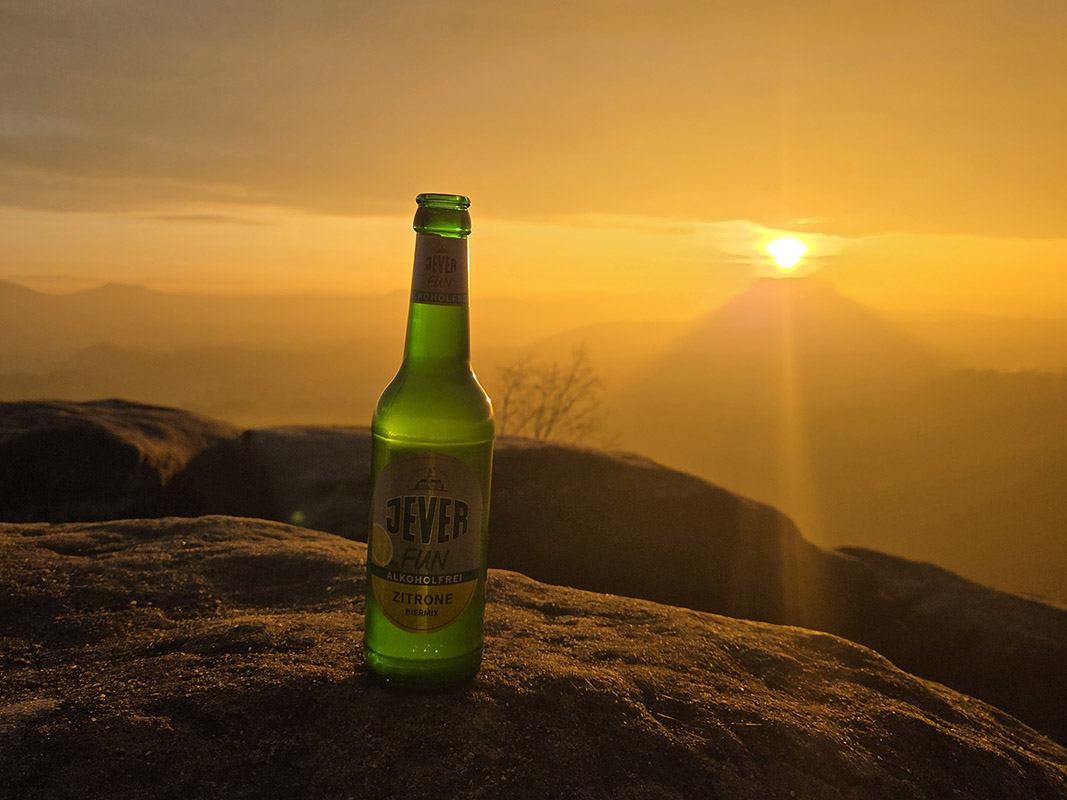 Eine Jever-Bierflasche steht auf der Spitze eines Berges. Im Hintergrund sind Berge und ein orangefarbener Himmel.