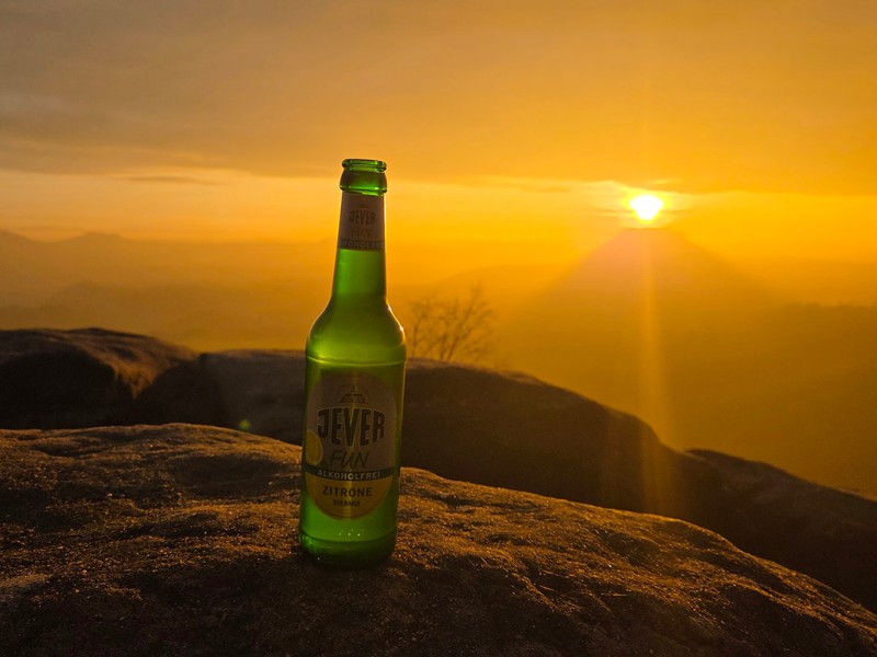 Eine Jever-Bierflasche steht auf der Spitze eines Berges. Im Hintergrund sind Berge und ein orangefarbener Himmel.