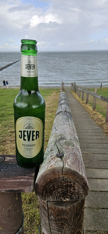Eine Weg mit einem Geländer jeweils rechts und links daneben führt runter zu einem Gewässer. Oben auf dem Hügel steht eine Jever-Bierflasche auf dem Geländer.
