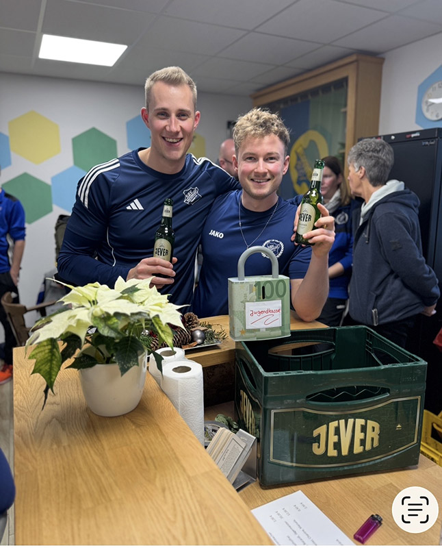 Zwei Männer in Sportkleidung stehen lächelnd nebeneinander hinter einem Tresen. Beide halten jeweils eine Jever-Bierflasche vor sich.
