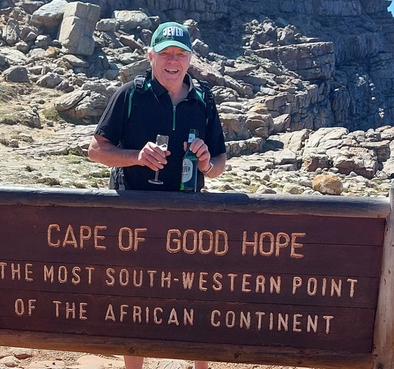 Ein Mann, der eine Jever-Mütze trägt, hält eine Jever-Bierflasche und ein Glas mit Bier in beiden Händen. Vor ihm steht ein Schild auf Englisch mit der Aufschrift Kap der Guten Hoffnung, der südwestlichste Punkt des afrikanischen Kontinents.