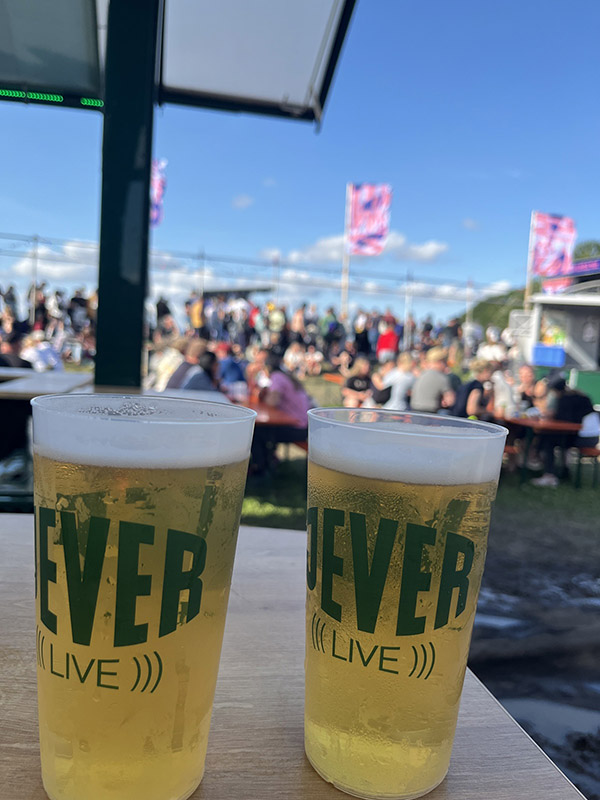 Zwei Becher mit der Aufschrift Jever live stehen nebeneinander auf einem Tisch. Im Hintergrund sitzen zahlreiche Personen auf Bänken mit Tischen von Bierzeltgarnituren.