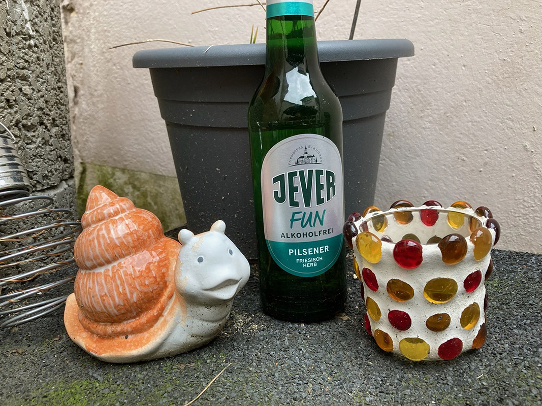 Eine Jever-Bierflasche steht vor einem Blumentopf. Links vom Glas steht eine dekorative Schnecke und rechts davon ein Glas für Teelichter.