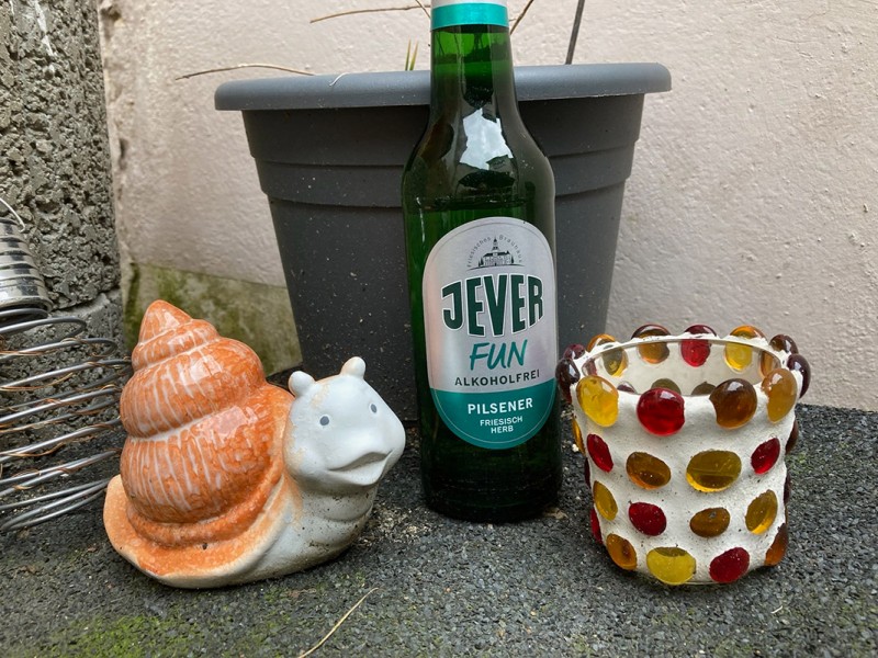 Eine Jever-Bierflasche steht vor einem Blumentopf. Links vom Glas steht eine dekorative Schnecke und rechts davon ein Glas für Teelichter.