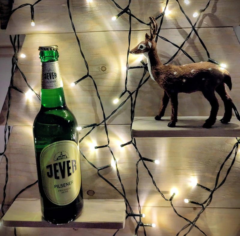 Auf einem Wandregal steht eine Jever-Bierflasche. Rechts darüber steht eine kleine Hirschfigur auf einem anderen Wandregal. Beides ist von einer Lichterkette umgeben.