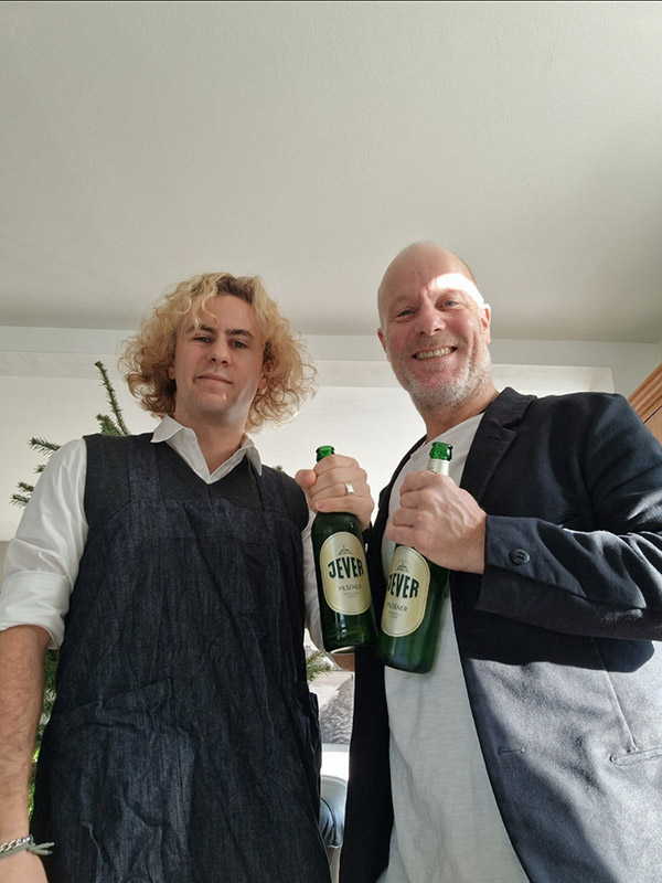 Zwei Männer stehen in einem Zimmer vor einem ungeschmückten Tannenbaum. Beide Männer halten jeweils eine Jever-Bierflasche vor sich.