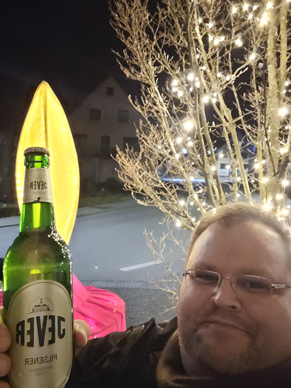 Ein Mann hält ene Jever-Bierflasche neben sein Gesicht. Hinter ihm ist ein blätterloser Baum mit einer Lichterkette.
