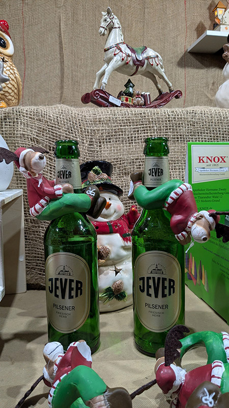 Zwei kleine Rentierfiguren mit Weihnachtskleidung hängen am Flaschenhals von Jever-Bierflaschen. Um sie herum sind weitere weihnachtliche Dokorationsartikel verteilt.