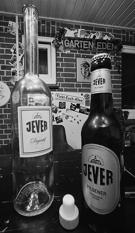 Eine Jever-Bierflasche und eine Flasche Jever Digestif stehen nebeneinander auf einem Tisch. Im Hintergrund hängen an der Wand zahlreiche Postkarten.