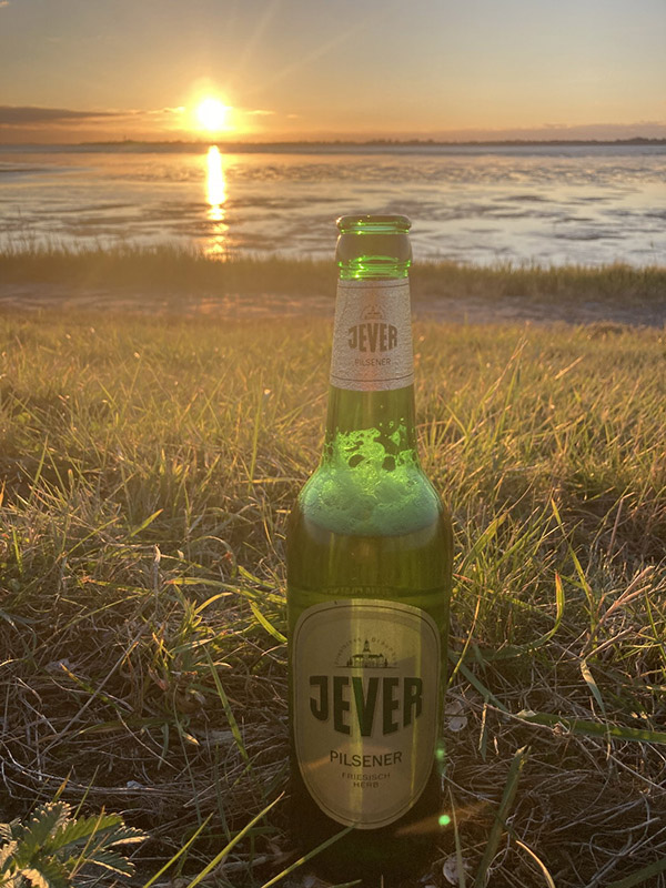Eine Jever-Bierflasche steht auf einer Wiese. Im Hintergrund ist ein Gewässer und die Sonne färbt den Himmel orange.