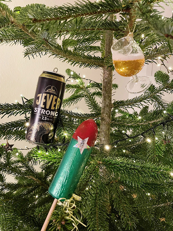 An einem Tannenbaum mit einer Lichterkette hängt ein Glas mit Bier, eine Jever-Bierflasche und eine Rakete.