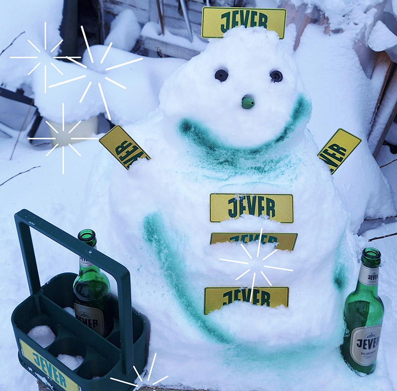 Ein Schneemann hat mehrere Jever-Schilder als Arme, Knöpfe und Hut. Links von ihm ist ein Jever-Bierflaschenträger und rechts eine Jever-Bierflasche.
