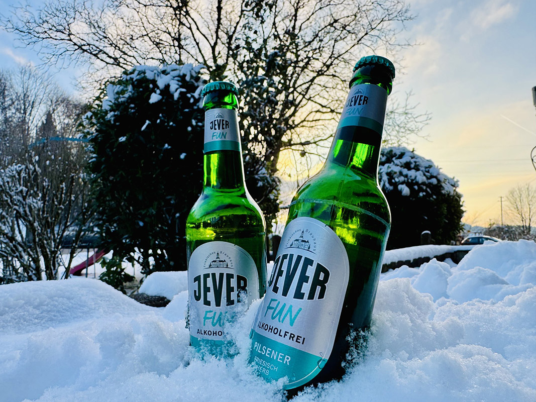 Zwei Jever-Bierflaschen stecken nebeneinander im Schnee. Im Hintergrund sind mehrere verschneite Bäume.