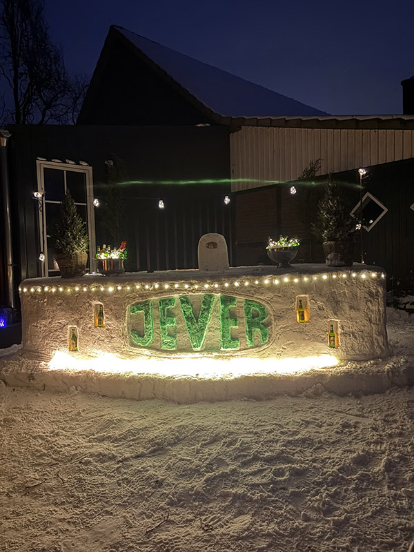 Ein Tresen aus Schnee mit beleuchtetem Jever-Schriftzug und mehreren eingebauten Bierflaschen steht in einem Garten.
