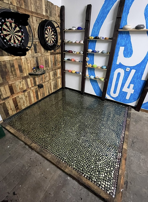 An einer Wand hängen zwei Dartboards nebeneinander. Auf dem Boden davor ist ein Rechteck aus zahlreichen Kronkorken.