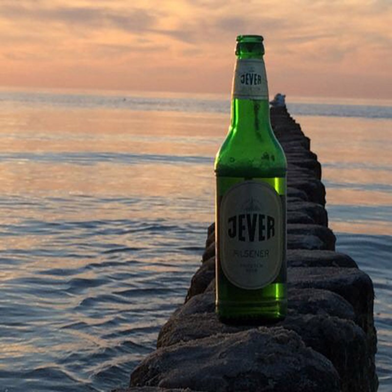 Eine Jever-Bierflasche steht auf einem von Wasser umgebenen Steinweg.